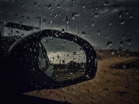 Close-up of a car side mirror with raindrops, capturing a wet and stormy atmosphere.